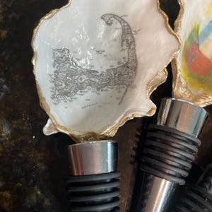 May include: A white oyster shell bottle stopper with a gold rim and a map of Cape Cod. The stopper has a silver metal base and a black rubber seal. The product is a decorative item.