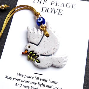 May include: A white ceramic dove ornament with black speckles, holding a green olive branch. It hangs from a gold cord with a blue evil eye bead. The display card reads: THE PEACE DOVE, May peace fill your home, May your heart stay light and protected, And may kindness always find its way to you, NAZAR LUCK DESIGN.