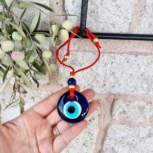 Puede incluir: Un amuleto de vidrio azul con forma de ojo turco, con un diseño de ojo blanco y azul, suspendido de un cordón rojo con cuentas doradas. El amuleto se muestra frente a una pared de ladrillos, con ramas de olivo en el fondo.
