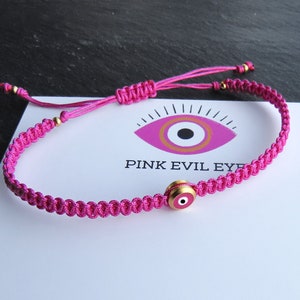 Puede incluir: Pulsera de cuerda rosa con un dije de ojo turco dorado. El dije tiene un diseño rosa, blanco y dorado. La pulsera está atada con un nudo y tiene una pequeña cuenta dorada al final.
