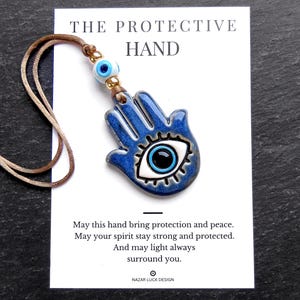 May include: A blue ceramic hamsa hand pendant with a central eye motif, attached to a brown cord with a small blue evil eye bead. The pendant is displayed on a white card featuring the text THE PROTECTIVE HAND and a printed blessing message.