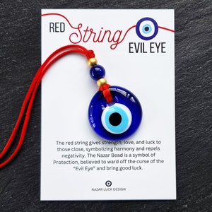 Può includere: Cordoncino rosso con un ciondolo di occhio turco blu e bianco. Il ciondolo è rotondo e ha una perla dorata in cima. Il cordoncino è annodato in fondo. La carta recita "RED String EVIL EYE" e spiega il significato del ciondolo di occhio turco.