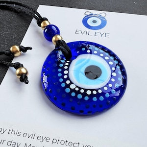 May include: A blue glass evil eye pendant with white and blue dots, on a black cord with gold beads. The pendant is round and has a white and blue eye design. The text "Evil Eye" is written on a white card behind the pendant.