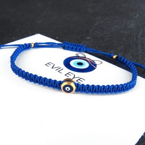 Puede incluir: Pulsera de cordón trenzado azul con un dije de ojo turco azul y dorado. El dije tiene un centro blanco y una pupilla negra. La pulsera está atada con un nudo y tiene cuentas doradas.