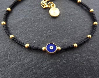 May include: A black beaded bracelet with gold beads and a blue evil eye charm. The bracelet has a gold chain with a small gold disc charm.