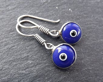 May include: A pair of silver-toned earrings featuring a blue glass eye design. Each earring has a round, cobalt blue glass cabochon with a white and black center, set in a silver-colored metal frame. The ear wires are a simple hook style.