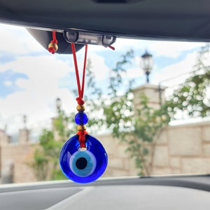 May include: A blue and white evil eye charm hanging from a red string with gold beads. The charm is hanging from the rearview mirror of a car.
