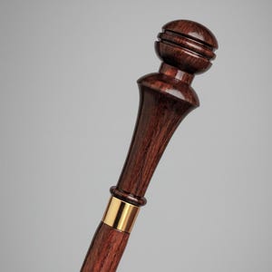 Può includere: Un bastone da passeggio in legno marrone scuro con un design decorativo. Il bastone presenta una parte superiore arrotondata con scanalature orizzontali, una fascia color oro e dettagli intagliati lungo l'asta. La venatura del legno è visibile.