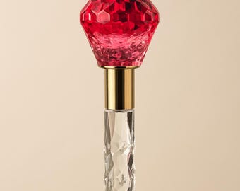 May include: A decorative bottle stopper with a faceted, ruby-red glass top. The stopper has a gold-colored metal band and a clear, diamond-cut glass stem. The design is elegant and suitable for wine bottles.