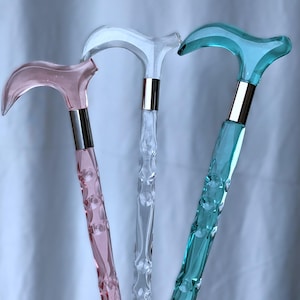 Puede incluir: Tres bastones con mangos curvos, que presentan un eje translúcido de color rosa, transparente y verde azulado. Cada bastón tiene una superficie texturizada y facetada, con un collar de metal plateado que conecta el mango con el cuerpo.
