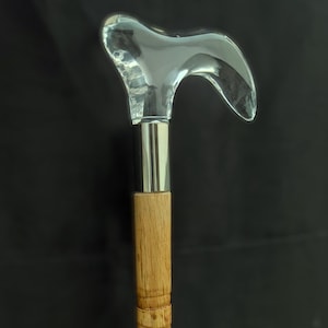 Op de afbeelding: Wandelstok met een heldere, gebogen glazen handgreep en een zilverkleurige metalen kraag. De houten schacht heeft gesneden diamantpatronen en een natuurlijke houten afwerking. De stok is ontworpen voor ondersteuning en stijl.