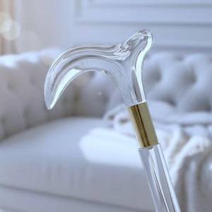 Könnte beinhalten: Ein klarer, gebogener Gehstock mit einem goldfarbenen Band. Der Griff ist transparent und ergonomisch geformt. Das Design des Gehstocks ist elegant und modern, geeignet für alle, die Stil und Unterstützung suchen.