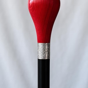 Puede incluir: Bastón de madera con un mango rojo vibrante y un eje negro. El mango tiene forma de lágrima y muestra la veta de la madera. Una banda plateada con un diseño ornamentado une el mango al eje.