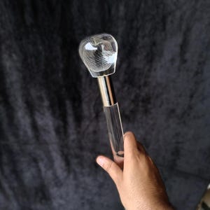 Puede incluir: Un bastón de acrílico transparente con un mango liso y redondeado que presenta un patrón de rejilla grabado en su interior. El mango está unido al eje transparente mediante un collar de metal plateado pulido, creando un apoyo de movilidad.