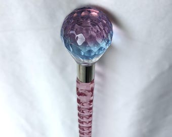 May include: A decorative walking cane with a faceted, spherical top that transitions from blue to purple. The shaft is pink with a textured, spiral design, and a silver metal collar connects the top and shaft.