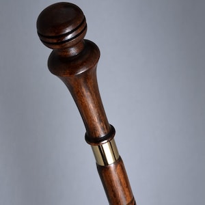 May include: A dark brown wooden walking cane with a rounded top and a brass band. The cane has a carved spiral design on the lower portion. The top has two carved rings. The cane is angled towards the right.
