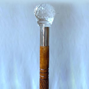 Puede incluir: Un bastón con un mango de bola de cristal transparente y facetada. El eje es de madera con una banda de metal plateado y un patrón tallado cerca de la parte inferior. El bastón mide aproximadamente 91 cm de largo.