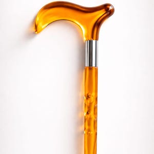 Puede incluir: Bastón de color ámbar con un mango curvo y un eje texturizado. El bastón tiene una banda de metal plateado donde el mango se une al eje. El bastón está sobre un fondo blanco. El texto "Smart Walk STORE" está en la parte superior.