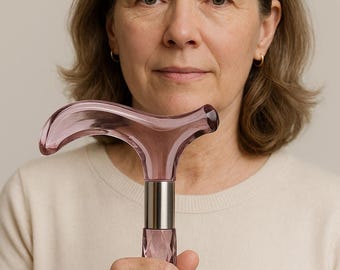May include: A pink, decorative walking cane with a faceted design. The handle is curved and the shaft is a matching pink color. The cane has a silver metal collar. The cane is held by a person wearing a cream-colored sweater.