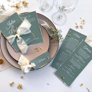 Könnte beinhalten: Hochzeitspapeterie-Set mit salbeigrünen Karten, weißer Schrift und zerrissenen Kanten. Enthält Einladung, Menü und Tischkarte, mit weißem Band, Blattgold und einem rosafarbenen Teller. Zwei Weingläser sind sichtbar.