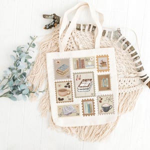 K&ouml;nnte beinhalten: Cremefarbene Tragetasche mit einer Reihe von Briefmarkenmotiven, die B&uuml;cher und Kaffeetassen zeigen. Die Tasche hat cremefarbene Griffe und ist vor einem wei&szlig;en Holzuntergrund mit dekorativen Elementen platziert.
