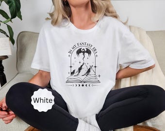 Könnte beinhalten: Weißes T-Shirt mit einem schwarzen Grafikdesign mit einem Drachen, Bergen und dem Text "IN MY FANTASY ERA". Das Design befindet sich in einem bogenförmigen Rahmen über einem offenen Buch mit Mondphasen. Das Shirt ist ein lässiges Kleidungsstück.