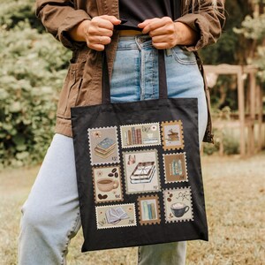 K&ouml;nnte beinhalten: Schwarze Tragetasche mit einer Collage von Briefmarken-Designs. Die Briefmarken zeigen B&uuml;cher, Kaffeetassen und andere literarische und Kaffee-bezogene Bilder. Die Tasche wird von einer Person gehalten, die eine braune Jacke und blaue Jeans tr&auml;gt.