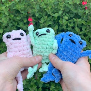 May include: Three handmade crochet frog toys in pink, green, and blue. Each frog has black button eyes and a stitched mouth. The frogs are held up against a background of green foliage.
