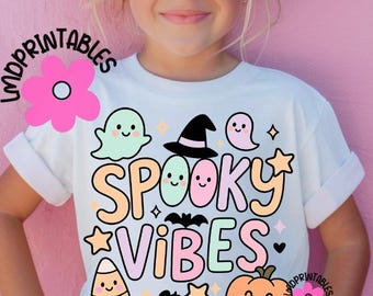 以下が含まれることがあります： パステルカラーの「Spooky Vibes」の文字が入った白いTシャツ。漫画の幽霊、魔女の帽子、カボチャ、トウモロコシのキャンディーが描かれています。サングラスをかけた幼い子供が着ています。