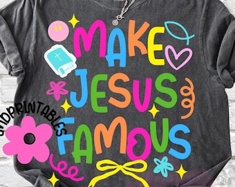 May include: Dark gray t-shirt with the phrase "MAKE JESUS FAMOUS" in colorful, playful lettering. The design includes a smiley face, a book, a heart, and other decorative elements. A silver necklace is visible.