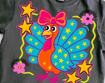 May include: Dark gray t-shirt featuring a colorful cartoon turkey with a pink bow, blue and yellow feathers, and yellow stars. The design includes the text "MDPRINTABLES" and is surrounded by yellow stars and pink flowers.