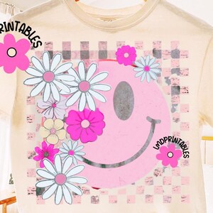 Op de afbeelding: Cr&egrave;mekleurig T-shirt met een grote roze smiley met een madeliefjesontwerp en een geblokte achtergrond. Het shirt heeft de tekst "LMDPrintables" erop. Het ontwerp bevat roze, wit en lichtblauwe kleuren.
