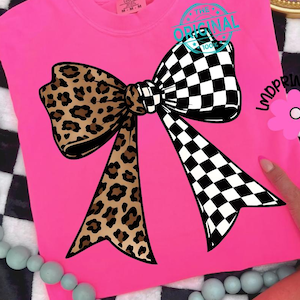 May include: A bright pink t-shirt featuring a large bow design. The bow is split, with a leopard print on the left and a black and white checkered pattern on the right. The text "THE ORIGINAL 100%" is printed on the top right.