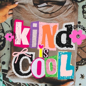 May include: A tan-colored t-shirt with the words "Kind is Cool" in a colorful, collage-style design. The text features various fonts and colors, including pink, orange, black, and blue. The shirt is held by hands.