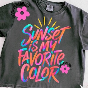 May include: Dark gray t-shirt with the text "Sunset is my Favorite Color" in vibrant, multi-colored lettering. The design includes a sun graphic and is accented with pink flower graphics.