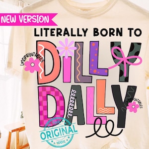 May include: Cream-colored t-shirt with the phrase "LITERALLY BORN TO DILLY DALLY" in colorful, playful lettering. The design includes floral accents and a pink bow. The shirt has a "NEW VERSION" banner and "ORIGINAL" stamp.