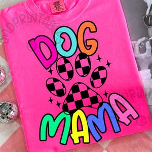 May include: Bright pink crew neck t-shirt with the words "DOG MAMA" in colorful, outlined letters. A paw print design with a checkered pattern is featured in the center. The shirt is a Comfort Colors brand.