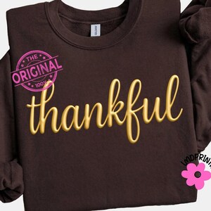 May include: A dark brown sweatshirt with the word "thankful" in gold script across the front. The text is embroidered. A pink stamp that says "The Original 100%" is in the upper left corner. A pink flower with the text "LMDRINTABLES" is in the lower right corner.