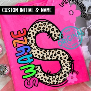 May include: A vibrant pink t-shirt featuring the word "SWAYZE" in rainbow hues and a leopard print "S" design. The text "CUSTOM INITIAL & NAME" is above the design. The shirt is displayed on a pink and white checkered surface.