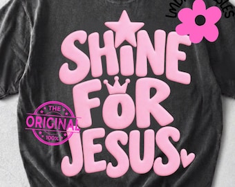 May include: A black short-sleeve t-shirt featuring the pink text SHINE FOR JESUS in a rounded, puffy font. The design includes a pink star above the text, a small crown above the letter O, and a small heart to the right of the word JESUS.