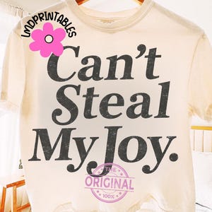 May include: Cream-colored t-shirt with the phrase "Can't Steal My Joy" in dark gray, distressed lettering. The shirt also features a pink flower graphic and a circular stamp that reads "The Original 100%."
