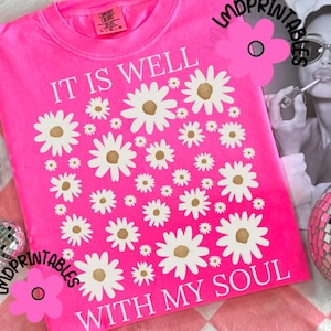 May include: A bright pink sweatshirt with the text "IT IS WELL WITH MY SOUL" and a daisy pattern. The Comfort Colors brand tag is visible. The image includes a newspaper clipping and disco ball.