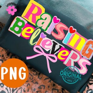 May include: A dark teal t-shirt with the words "Raising Believers" in colorful, cut-out style letters. A pink bow is below the text. The image also includes a PNG logo and a "Original 100%" stamp.