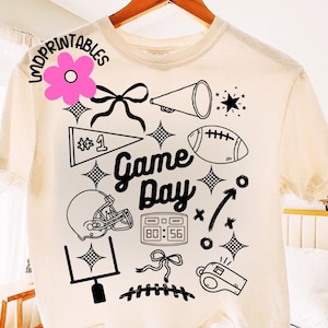 May include: Cream-colored t-shirt with a black graphic design featuring football-themed elements. The design includes a football, helmet, megaphone, and the words "Game Day". The shirt also has a score display and a whistle.
