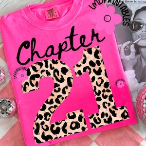 May include: A bright pink sweatshirt with the words "Chapter 21" in black script and leopard print. The sweatshirt is a Comfort Colors brand. The background includes a newspaper clipping and a disco ball.