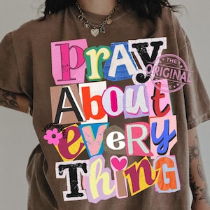 May include: Brown t-shirt with the phrase "Pray About Everything" in a colorful, cut-out style design. The text is arranged in a stacked format with various colors and fonts, creating a collage effect. A pink flower and heart are also included.