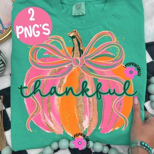 Peut inclure: Un t-shirt Comfort Colors turquoise avec un motif de citrouille color&eacute;e. La citrouille est orange et rose avec des paillettes dor&eacute;es et un grand n&oelig;ud rose. Le mot "thankful" est &eacute;crit en vert. Un cercle rose avec "2 PNG'S" est en haut &agrave; gauche.