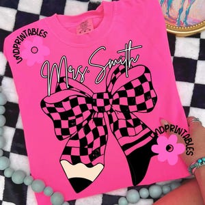 May include: A vibrant pink t-shirt featuring a large black and pink chequered bow design and a pencil. The name "Mrs. Smith" is written in cursive above the bow. The shirt has the words "Unprintables" and a pink flower graphic.