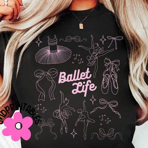 May include: Black t-shirt with a pink ballet-themed design. The design includes illustrations of a tutu, ballet shoes, bows, and dancers, with the words "Ballet Life" in a script font.