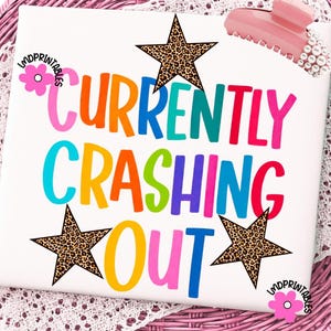 以下が含まれることがあります： カラフルな文字で「CURRENTLY CRASHING OUT」と書かれた白い正方形。ヒョウ柄の星がテキストの周りに配置されています。ピンク色のヘアクリップとパールの装飾が右上に見えます。